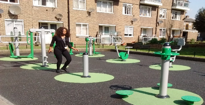 Young woman in black top & trousers walks across the new gym equipment in the courtyard of Caldwell gardens estate