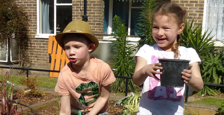 Boy in hat holding top of a fork and gril in pink & white t-shirt working on raised wooden flowerbed at community planting day Caldwell Gardens Spring 2017