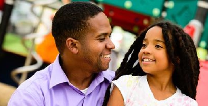 Man in a lilac shirt with a young girl, in a park playground looking at each other and smiling.