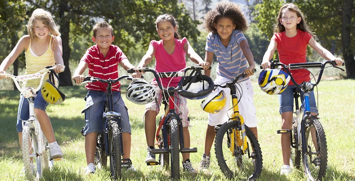 Left to right: blonde girl, yellow top; tan younger boy striped polo shirt; dark-haired girl; child with dark corkscrew hair ;; dark haired girl in red T all on bikes in a summer field - multicoloured safety helmets