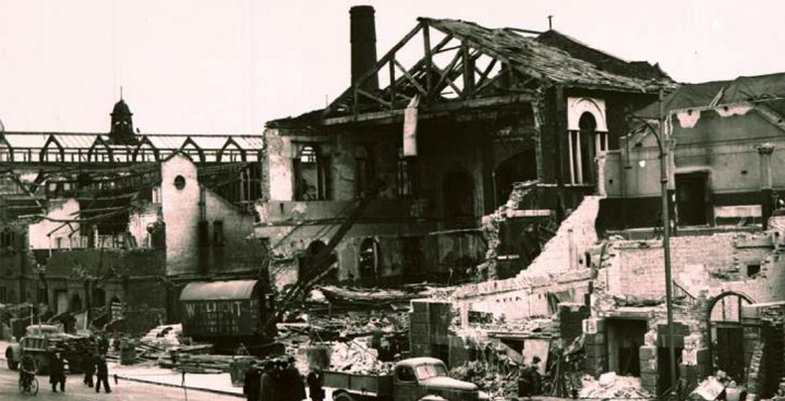 V2s in Lambeth talk. B&W photo of bombsite in Lambeth after a WW2 V2-bomb attack, Ruined house by metal railway bridge with horsedrawn and motorised vehicles in road outside and people in hats long coats