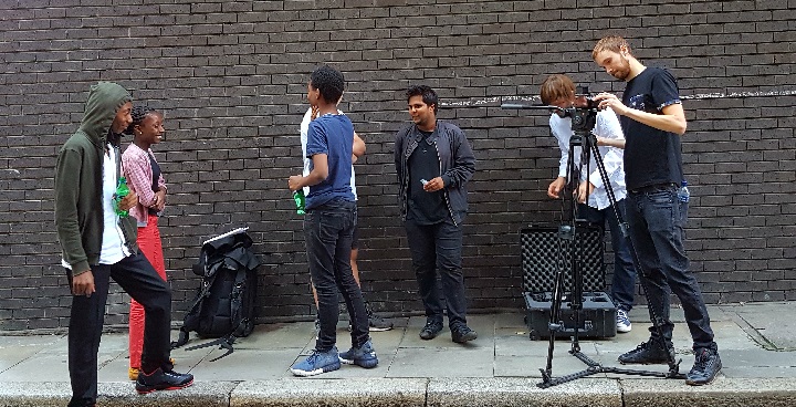 Creative Sparkworks film crew of young people learning to use cameras & film sound recording equipment on streets with black brick wall in background