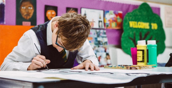Child sitting in a school classroom, bending over the desk as he paints.