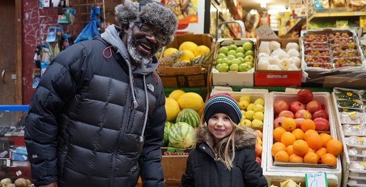 adult and child in winter clothes in fromt of shelves of fruit and veg in shop or covered market