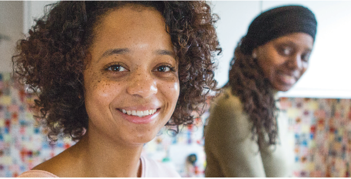 close up face of teenage girl smiling with light brown skin, curly hair & light pink lipstick; smiling adult woman in background in green top and headscarf