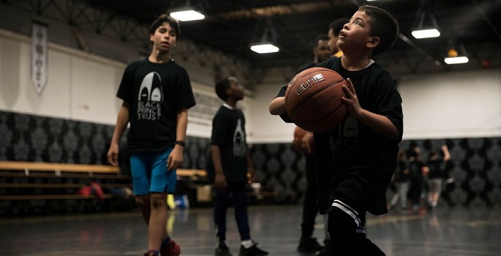 3 x pre-teen boys on Black Prince TRust t-shirts in action of the basketball court - boy in foreground crouching holding ball