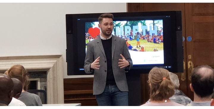 Cllr Matt Bennett in grey jacket and jeans speaks at Crowdfunding Event, Lambeth Town Hall Feb 2018 in front of a screen with the Lamveth Crowdfunding image of a crowd standing around life-size 'Lambeth' letters