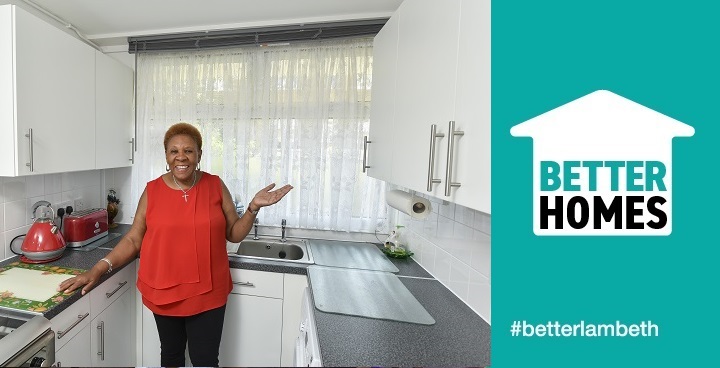 Short haired 50-something Caribbean woman in red frilly top and dark trousers Valencia points at new white cubloard doors and dark grey worktops in her newly updated kitchen July 2018