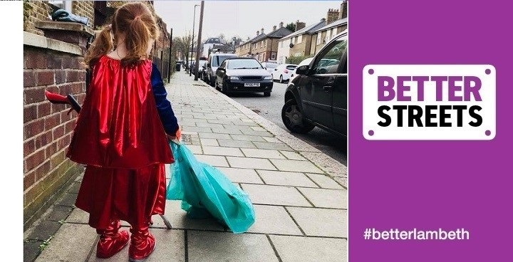 Pre-school age blonde girl with hair in bunches wearing a superman cape and outfit putting rubbish into a green bag on a Lambeth street (photographed from behind)