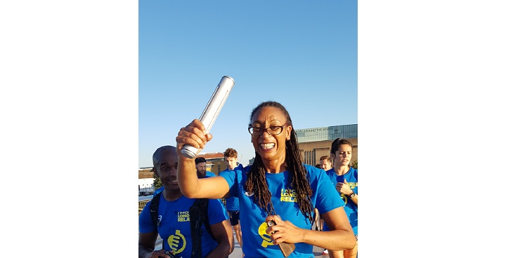 Runner from Transform wellness in blue lycra kit, glasses and long hair in braids holds the baton in the first 5k on a world record attempt for longest continuous run London JUly 2018