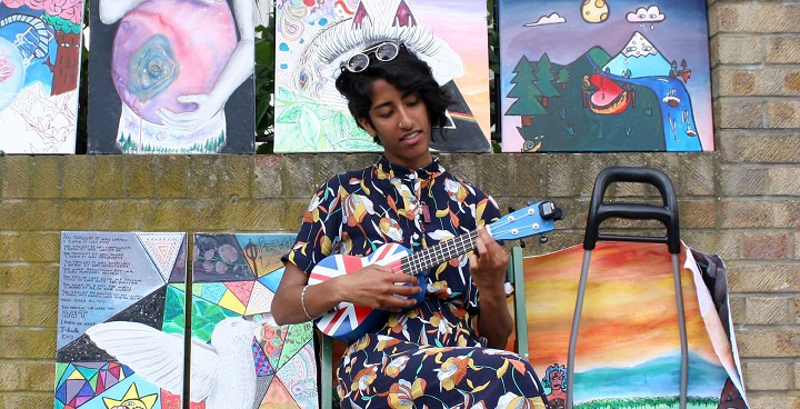 painter in floor length bold printed outfit plays union jack patterned ukelele in front of her paintings hung on the railings of Josephine Avenue SW2