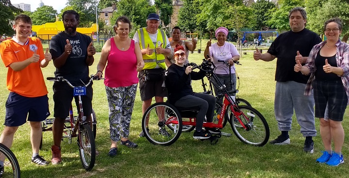 9 people of different ages including someone using adapated cycle give 'thumbs up' sign to Outdoor sport & exercise sessions for people with disabilities - part of a year-round programme for people & their carers