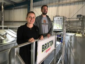Brixton Brewery co-founders Xochitl Benjamin and Jez Galaun standing in the brewery with large metal beer tanks in the background.
