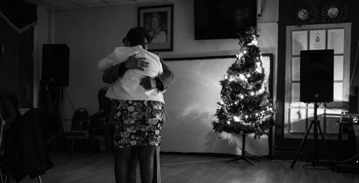 B&W photo of older Caribbean couple dancing in front of n Xmas tree - part of the 'Generation Windrsuh' photo exhibition on show in churches in Clapham & Angell Town in October & November