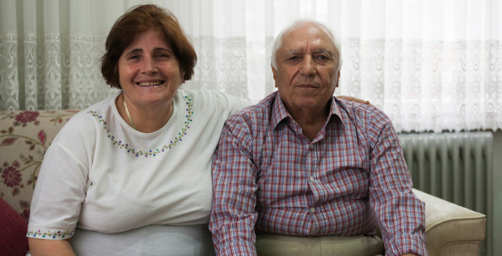 An older couple sat on a sofa smiling at the camera.