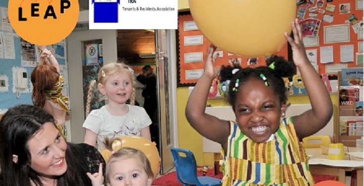 Foreground: toddler with big yellow balloon over her head; smiling babies & toddlers seated round her
