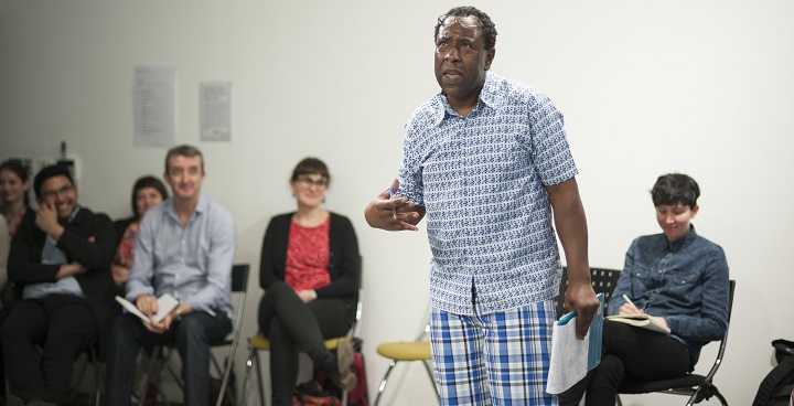 Actor Tony Cealy (pale blue shirt, check trousers, glasses on head) stands up with notes in his hand at a theatre workshop with seated adults