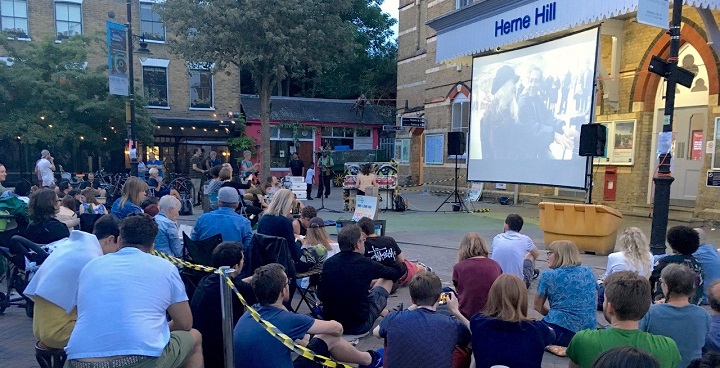 Herne Hill Free Film Festival - moviegoers outside Herne Hill station watching piano player on big screen