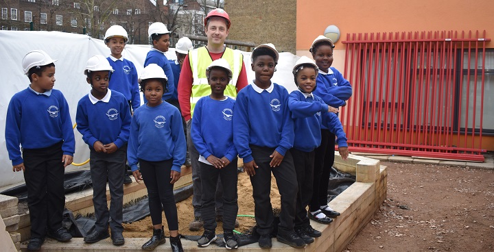 9 primary school children in blue schoool uniform jumpers, black trousers & white hard hats visit construction site of rebuilt Brixton childrens (early years) centre getting £1/2 million rebuild. Adult white male in hi-viz yellow & red safety helmet keeps an eye on their safety