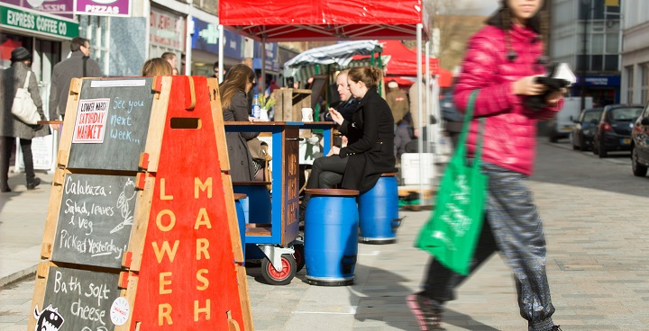 stallholders on lower marsh market by 'lower marsh' orange pyramid sign with chalkboard advertising farm cheeses; oriental woman in puffer coat walks past (out of focus) front right of the picture