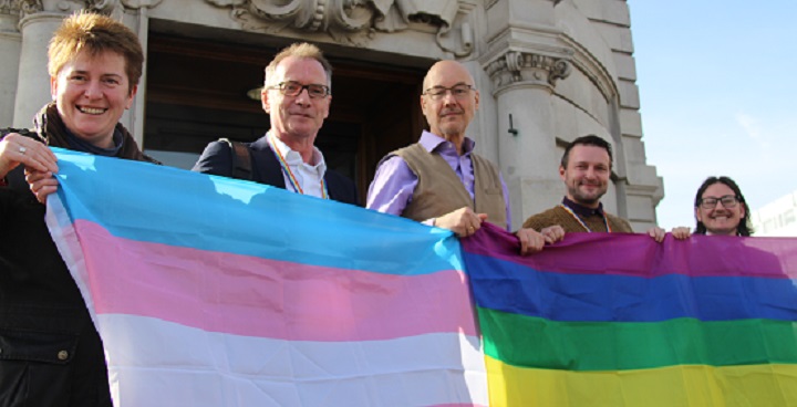 LGBT Foundation w/ Lambeth Council’s new Leader, the Chair of the CCG, the Council’s CEO, and the Strategic Director for Adults and Health draped in rainbow & trans flags on steps on the Town Hall l