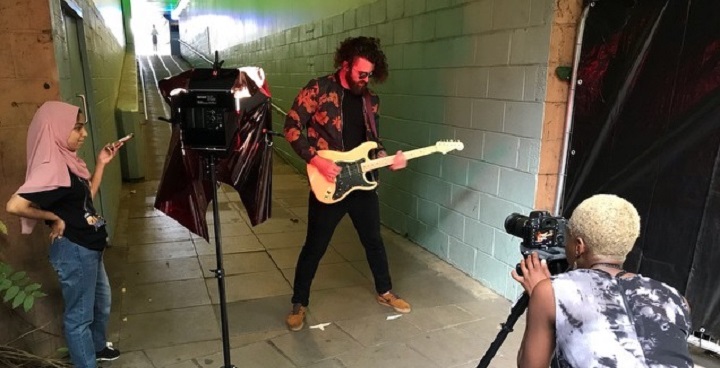 woman with dyed blonde hair (right) operate camera; women in hajib (left) directs a performance by young bearded man in red & black flowery shirt playing white electric guitar