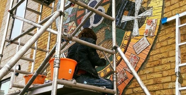 man in hoody sitting on scaffolding to install ceramics creating a mosaic of 'iconic places in Brixton' chosen/designed by SEND pupils at Lansdowne school