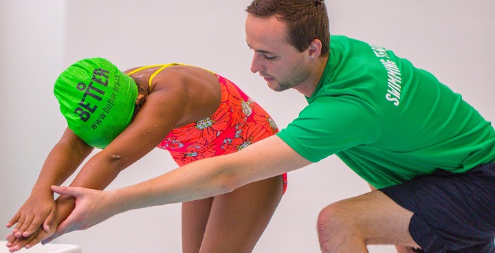 Child in green GLL better swimming cap and red 1-piece practices diving pose helped by swim teacher in same coloured green t-shirt