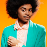 Woman creating her drag king persona - dressed as man with imperial beard and moustache, afro hair, green dinner jacket on peach background