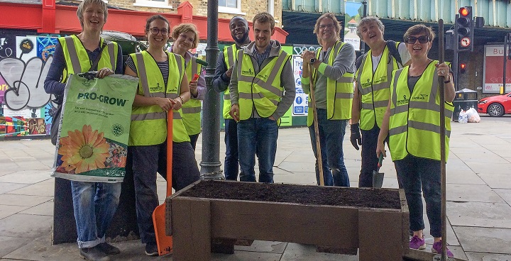 group of 6 volunteers in hi-viz ytellow vests lean on spoades to rest after filling planters with compost outside Herne Hill Station