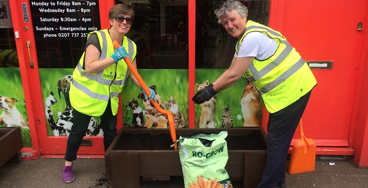 2 x women volunteers with red spades and yellow hi-viz vests fill a planter with compost made from recycled garden waste outside the pet shop opposite Herne Hill station
