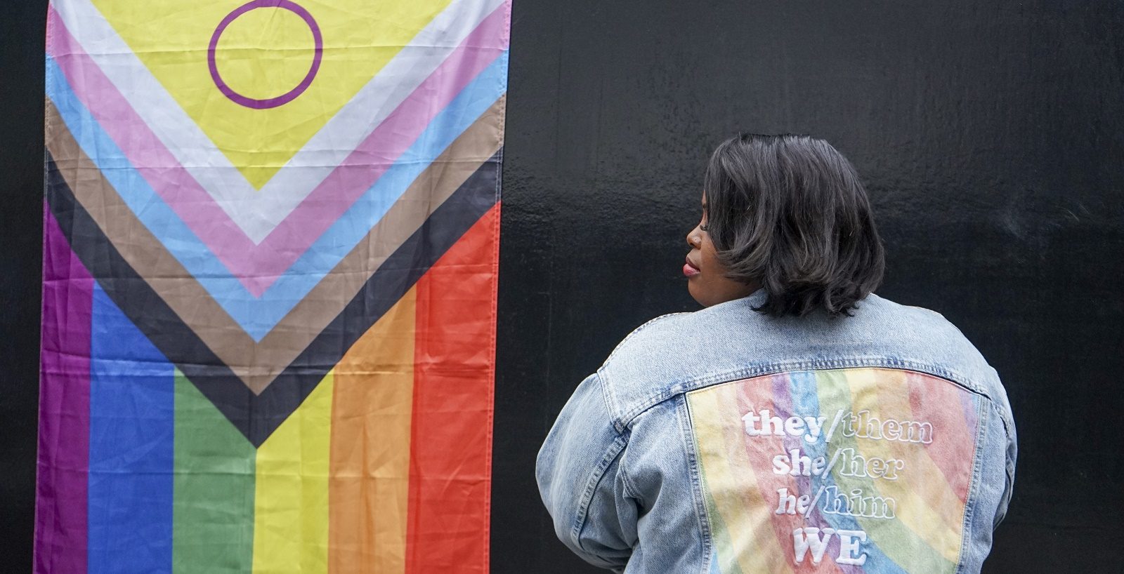 Poet Andreena Leeanne showing back of denim trucker jacket with rainbow flag embroidered with slogan they/them she/her he/him WE faces progress flag - photo Andreena Leanne