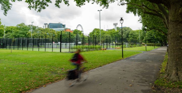 cyclist passing sports field in Lambeth park with London Eye in background