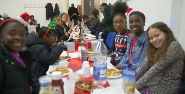 Group of boys and girls smiling at camera. Some are eating and wearing Christmas hats.