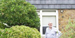 White male aged 70-80 standing in his front garden surrounded by shrubbery looking towards his roof.