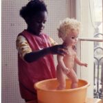 Photo of little black firl in pink dress washing a blonde baby doll (life sized) 1960s photo from Manning Studios