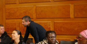 Man bends over to better hear what seated man is saying to him.