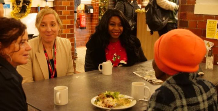 2 white women and a glamourous black lady in a red top sit opposite an elderly black woman wearing a woolly hat as she eats her lunch.