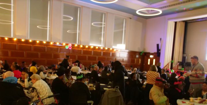 Large group of mostly older people sitting in a decorated hall, some wearing party hats.