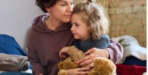 Woman sitting on bed with young girl clutching a teddy bear