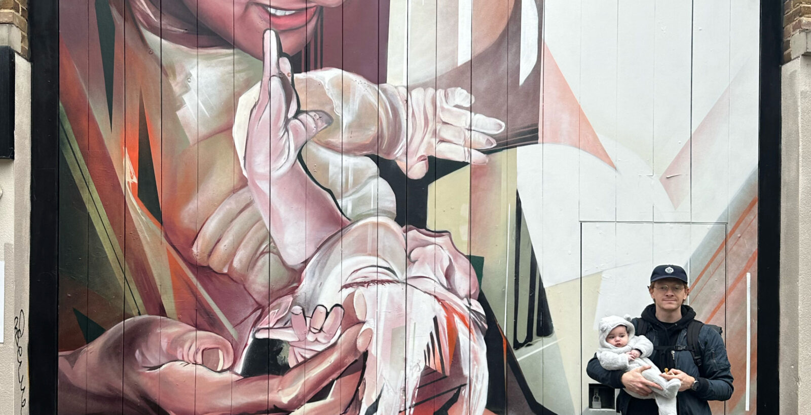 Artist Jake Attewell stands holding a baby in front of one of his large murals depicting a parent cradling a child in a blend of realism and abstract forms.