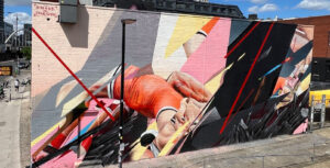 A large mural by artist Jake Attewell covers the side of a building in Stratford, combining realistic figures with bold abstract shapes and vibrant colours.