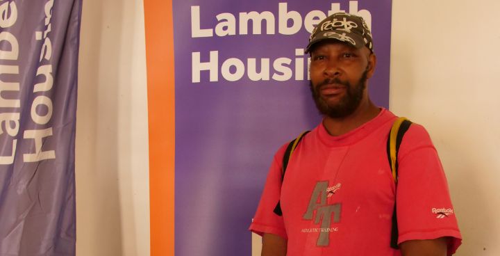 Man of Caribbean heritage wearing baseball cap and red tshirt stands in front of Lambeth Housing sign.